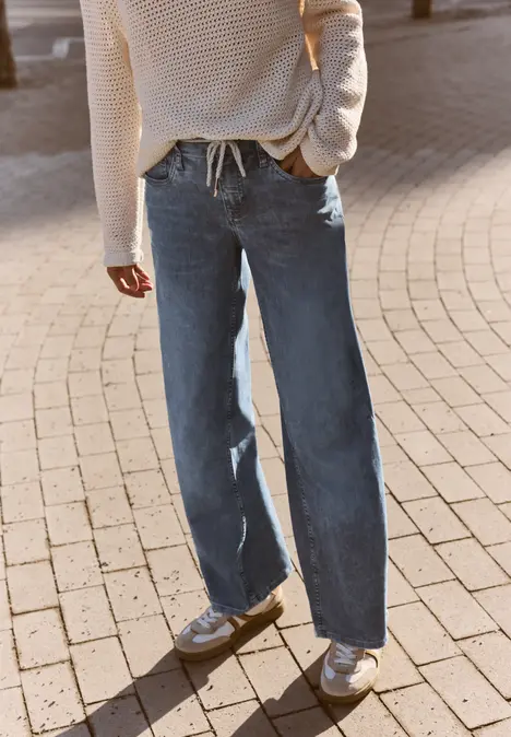 Persoon staat op een klinkerweg, draagt beige trui, wijde blauwe jeans en witte sneakers.