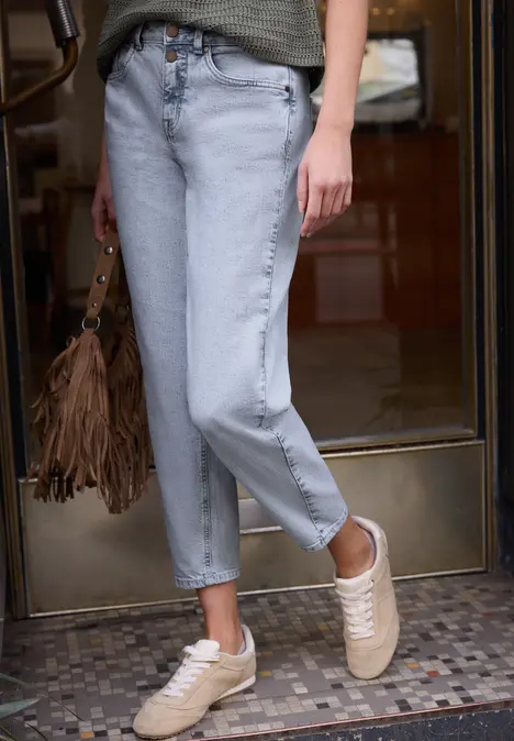 Een persoon draagt lichtblauwe cropped jeans, beige sneakers en een bruine franjes-tas voor een deur.