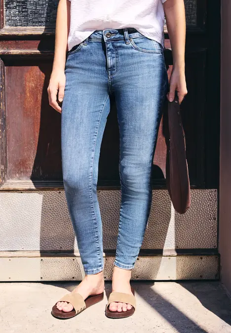 Eine Person trägt enge blaue Jeans und Sandalen, hält eine Tasche und steht vor einer Holztür.