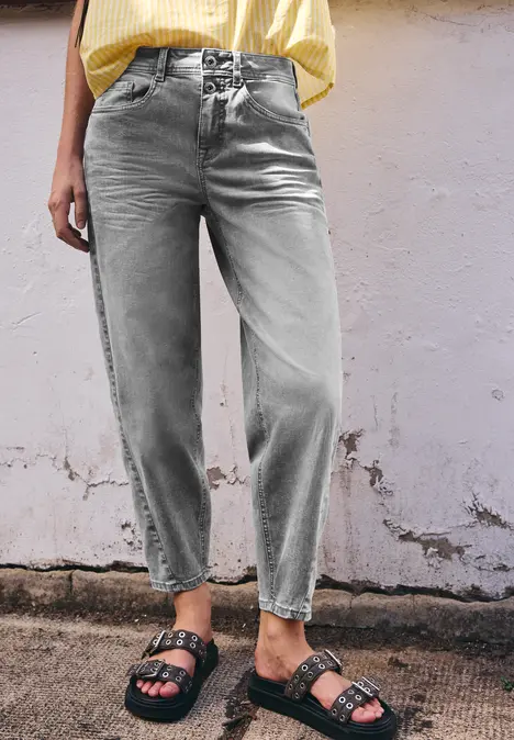Frau trägt weite, hellgraue Jeans und schwarze Sandalen vor einer abblätternden Wand.