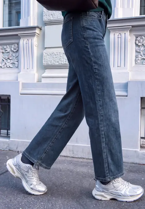 Beine in weiten Jeans und silbernen Sneakern beim Gehen vor einer Hausfassade.