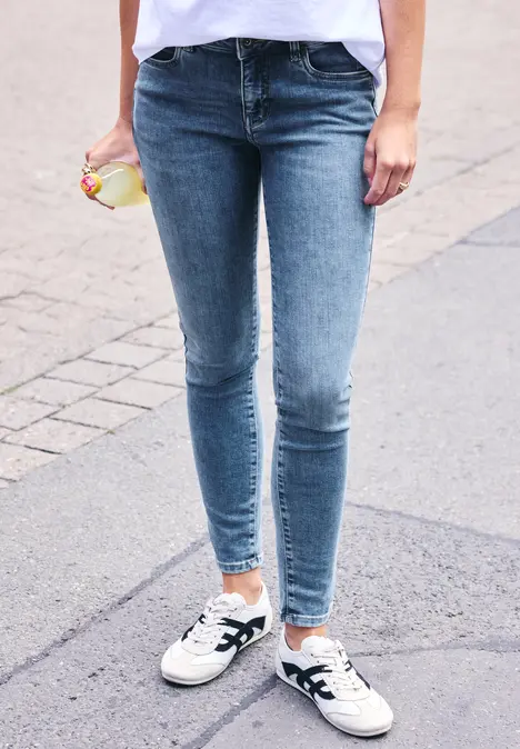 Person in engen Jeans und weißen Sneakers mit schwarzen Streifen läuft auf Bürgersteig und hält ein Getränk.