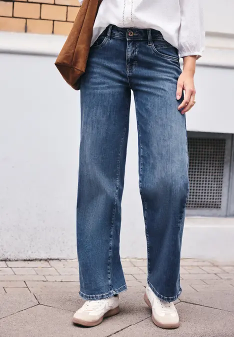 Eine Person trägt weite blaue Jeans, weiße Schuhe, weiße Bluse und trägt eine braune Tasche.
