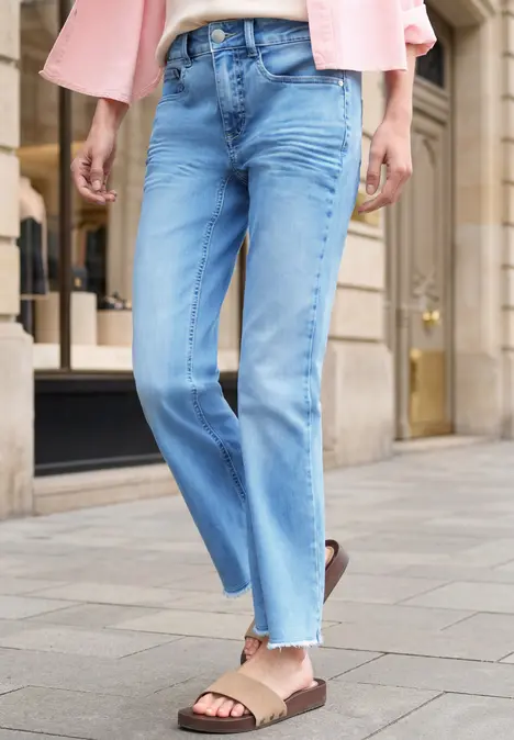 Vrouw in lichte, recht gesneden jeans met gefranjerde zoom en beige sandalen op de stoep.
