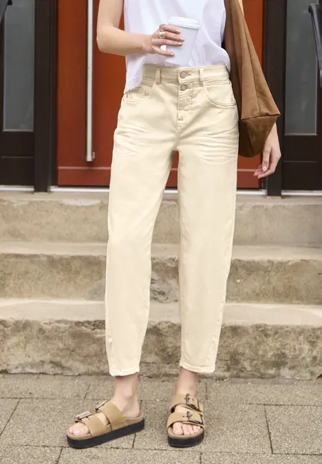 Persoon in beige broek en sandalen met stoffen tas voor een rode deur op een stoep.