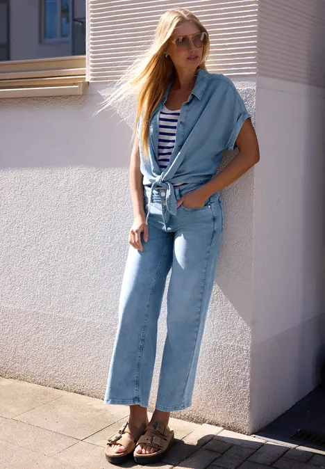 Junge Frau lehnt sich in hellblauer Denim-Culotte mit kurzen Ärmeln und gestreiftem Top an eine helle Hauswand.