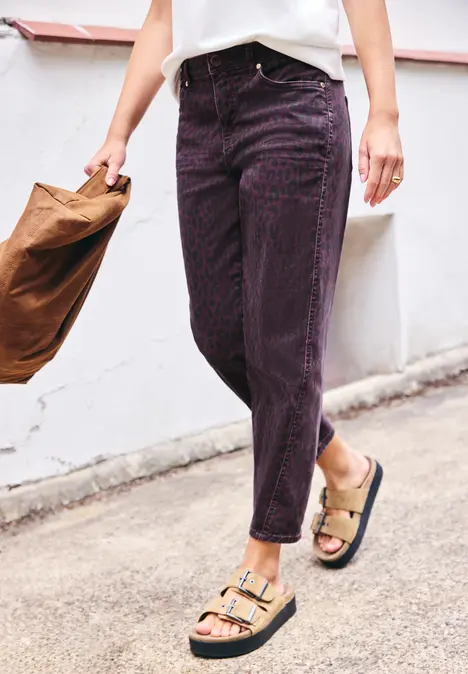 Eine Person in lila Jeans und beigen Sandalen mit Schnallen trägt eine braune Tasche.