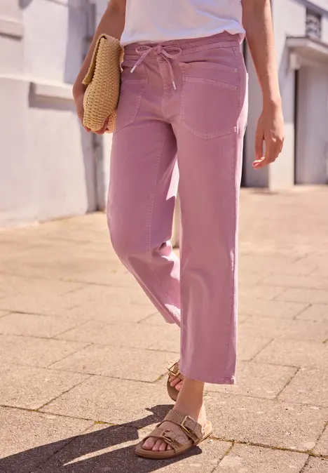 Vrouw loopt op een zonnig trottoir in losse, roze broek en sandalen, met strooien clutch in haar hand.