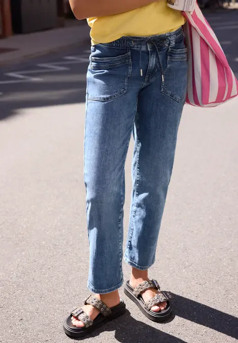 Ein Kind steht auf sonnigem Asphalt, trägt blaue Jeans, schwarze Sandalen und hält eine rosa-weiß gestreifte Tasche.