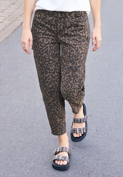 Eine Person geht auf grauem Asphalt, trägt braune, leopardgemusterte Hose und schwarze Sandalen mit silbernen Schnallen.