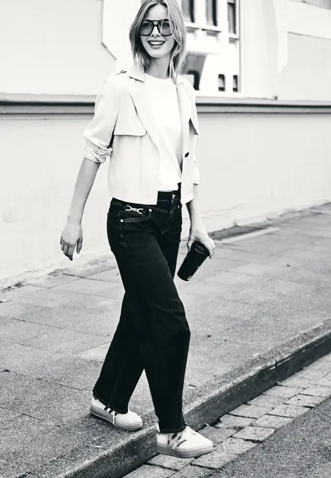Frau in Jeans,Sneakers,leichtem Mantel und Sonnenbrille spaziert lächelnd die Straße entlang,eine Clutch in der Hand.