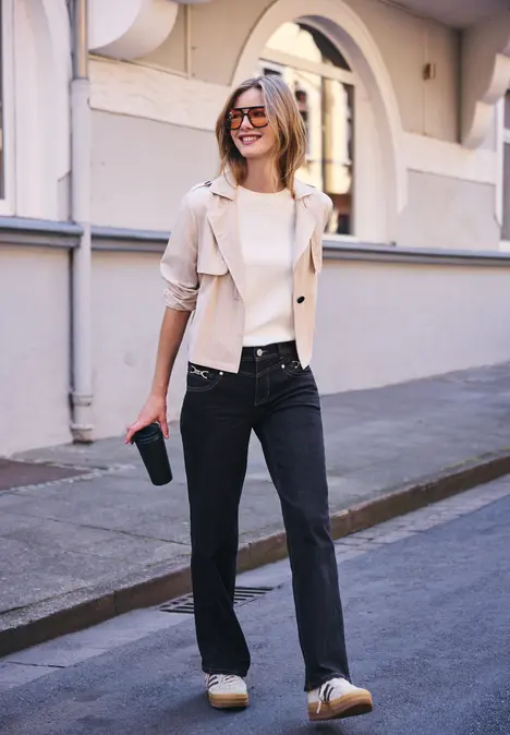 Vrouw met zonnebril, lichte blazer, zwarte jeans en laarzen, met coffee-to-go, glimlachend lopend op straat.