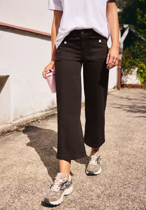 Person in weißem T-Shirt, schwarzen 7/8-Hosen und Sneakers mit rosa Becher auf dem Bürgersteig.
