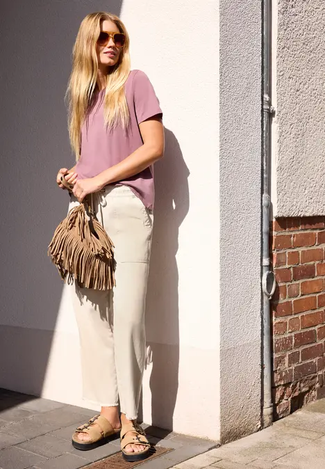 Junge Frau mit langen blonden Haaren, Sonnenbrille, rosa T-Shirt, hellen Hose und Fransentasche lehnt an Mauer.