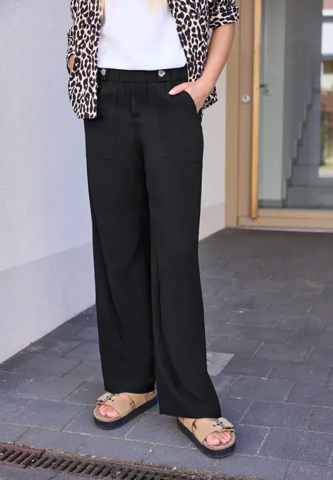 Frau in schwarzen, weiten Hosen, weißem T-Shirt, Leopardenjacke und beigen Sandalen steht vor Hauswand.
