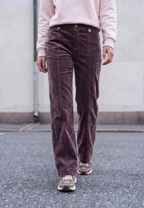 Personne de la taille aux pieds portant un pantalon velours côtelé violet et des baskets, marchant sur l'asphalte devant un mur.