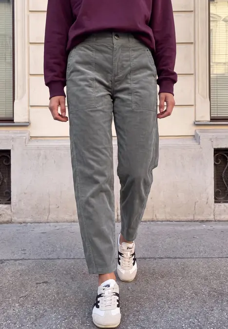 Personne en pantalon velours côtelé vert, baskets blanches à motif étoile et pull bordeaux, debout devant une façade.