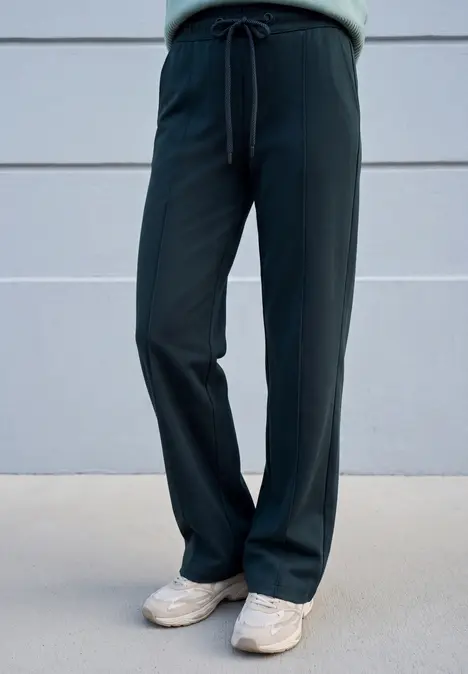 Schlanke Person trägt schwarze weite Hose mit Kordelzug und weiße Sneaker vor grauer Wand.
