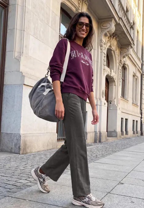 Junge Frau in lässigem Outfit und Rucksack spaziert an einer historischen Hausfassade entlang.