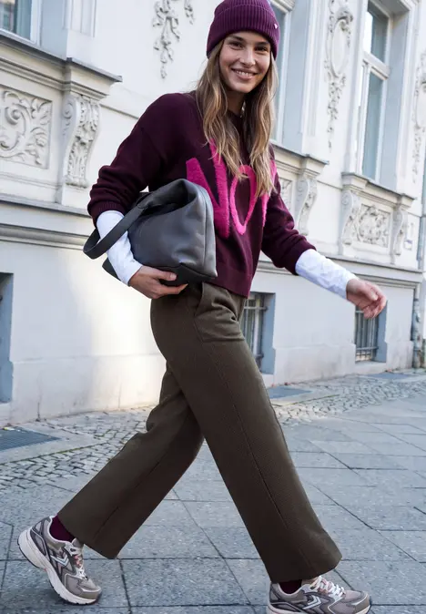 Een vrouw in winterkleding met muts en tas wandelt vrolijk over straat.