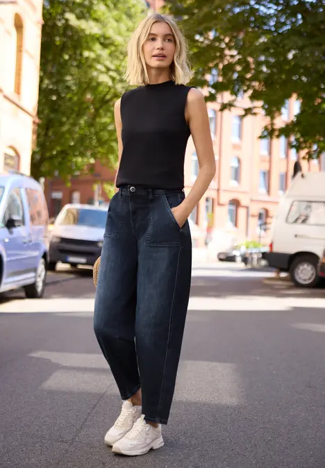 Eine Frau steht auf einer sonnigen Straße vor parkenden Autos, trägt ein schwarzes Top, weite Jeans und weiße Sneaker.