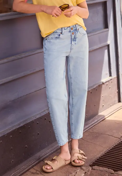 Persoon leunt tegen een muur, draagt een gele top, lichtblauwe high-waist spijkerbroek en beige sandalen.