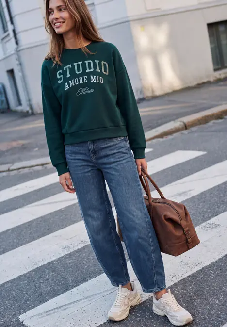 Een vrouw in een groen sweatshirt, blauwe jeans en witte sneakers loopt glimlachend over straat met een bruine tas.
