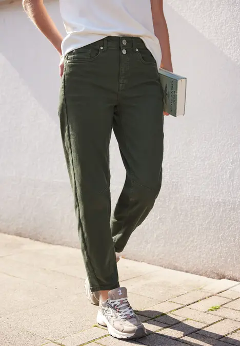 Personne debout sur le trottoir, porte un pantalon cargo vert olive, baskets, un livre à la main.