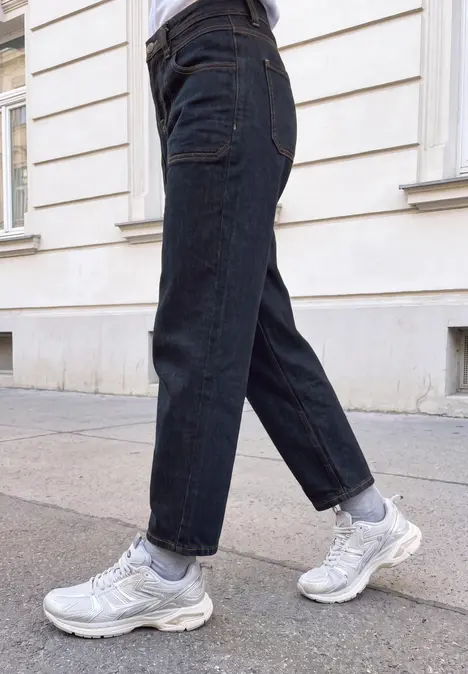 Eine Person läuft auf dem Gehweg in dunklen Jeans, weißen Socken und weißen Sneakern vor einer Hauswand.