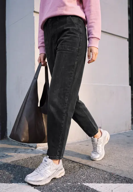 Person in schwarzer Cordhose, rosa Pullover und weißen Sneakern trägt schwarze Tasche und geht über Gehweg.