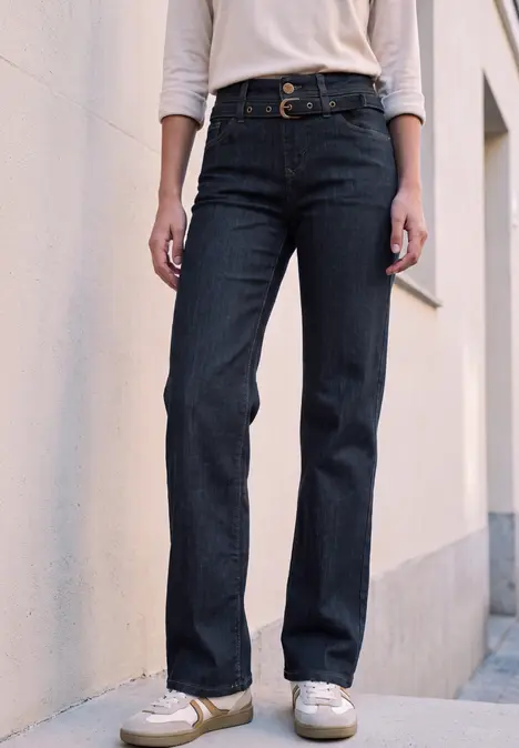 Personne porte un jean foncé, un haut blanc et des baskets blanches contre un mur.
