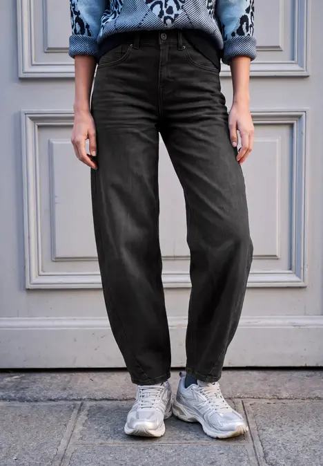 Person steht vor einer grauen Tür, trägt weite schwarze Hose und weiße Sneaker.