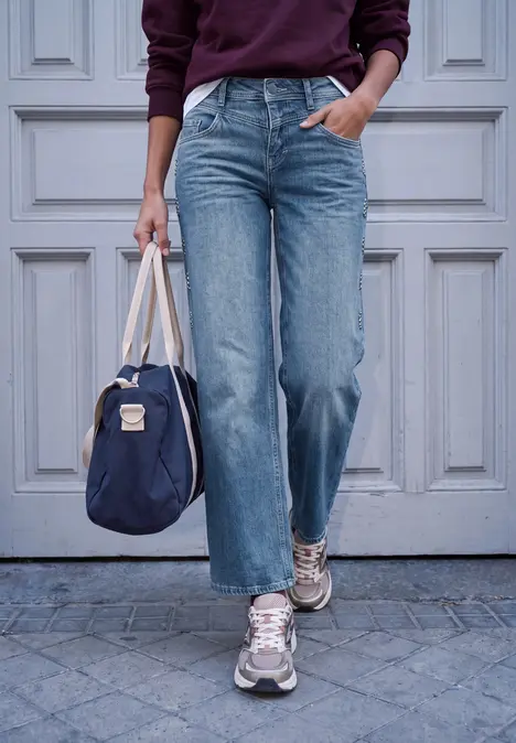 Person trägt weite blaue Jeans, Sneakers und hält eine dunkelblaue Tasche vor einer hellen Tür.