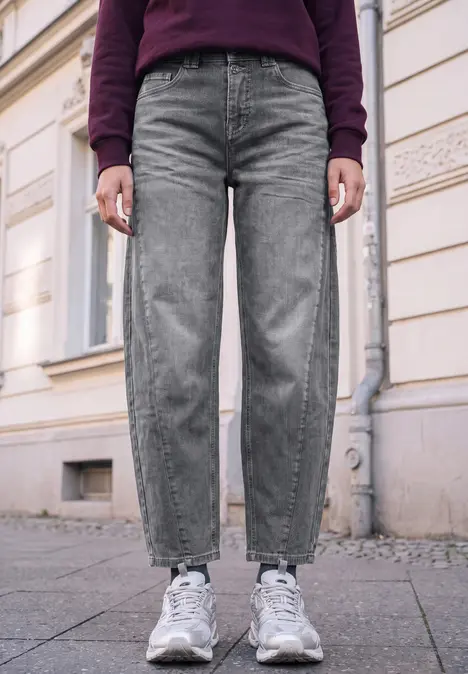 Unterkörper einer Person in grauen Jeans und weißen Sneakern auf Bürgersteig vor einer Hauswand.