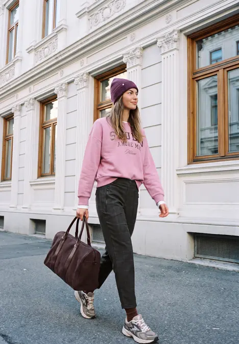 Junge Frau in pinkem Hoodie und Mütze spaziert mit Tasche vor klassischer Fassade.
