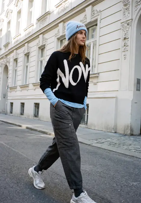 Frau in schwarzem 'NOW'-Pullover, blauem Rollkragen und Jogginghose mit weißen Sneakern vor elegantem Altbau.