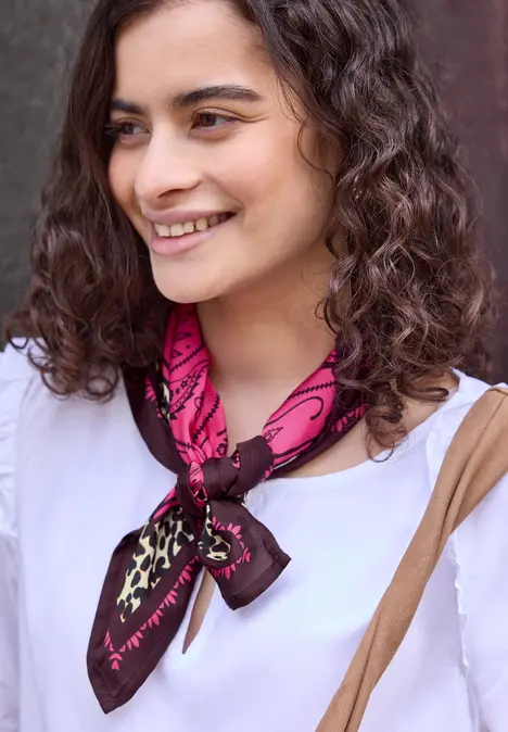 Person mit weißer Bluse, pink-braunem Halstuch und Umhängetasche, lockiges Haar, Gesicht unkenntlich gemacht