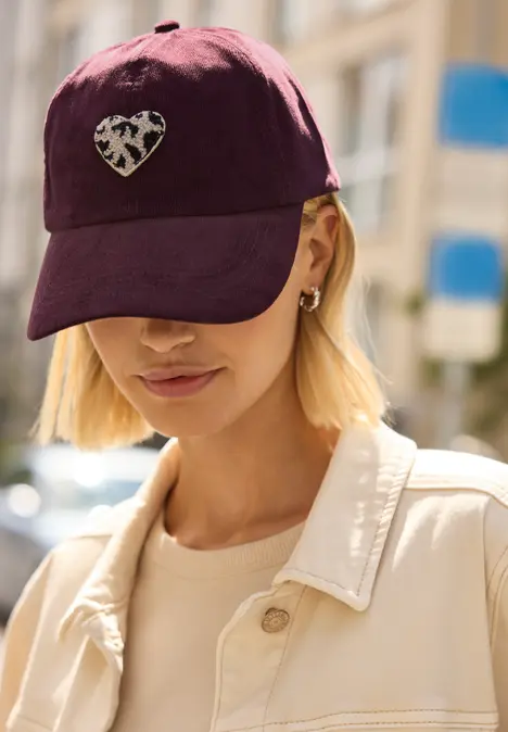 Person mit blonden Haaren trägt ein bordeauxfarbenes Cap mit Herzmotiv, goldene Ohrringe und helle Jacke.